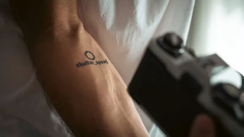 Arm with shutter_speed tattoo, analog camera blurred in the background
