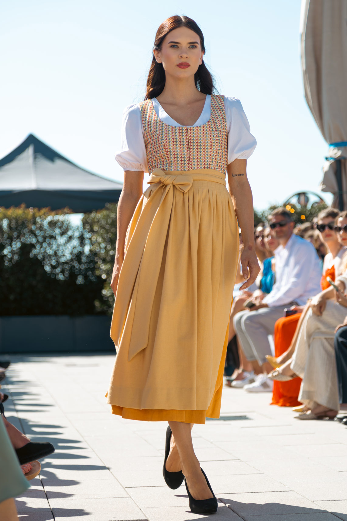 Model wearing a modern dirndl with a patterned bodice, white puff-sleeve blouse, and mustard-yellow skirt with a bow, walking an outdoor runway with the audience seated on both sides.