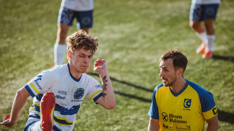 An opponent mid-kick with a scrunched-up face, the Gries captain beside him looking away