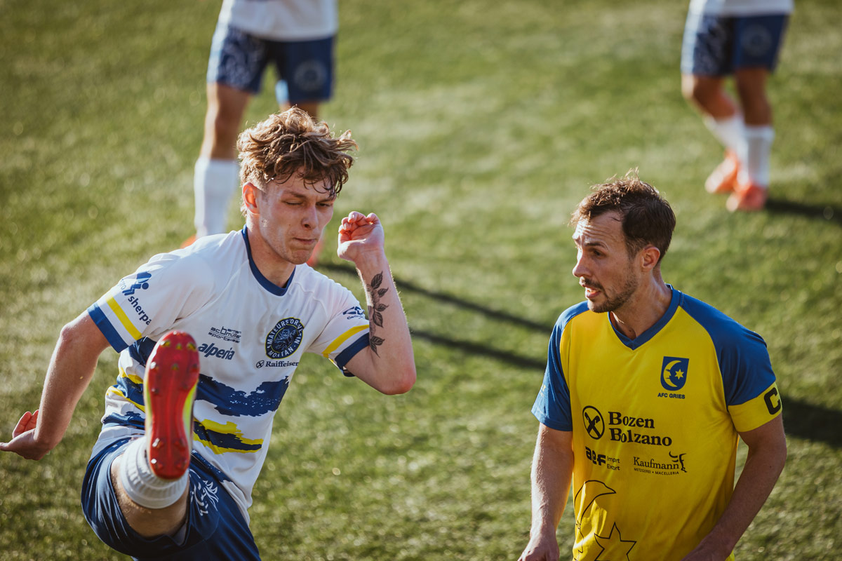 An opponent mid-kick with a scrunched-up face, the Gries captain beside him looking away