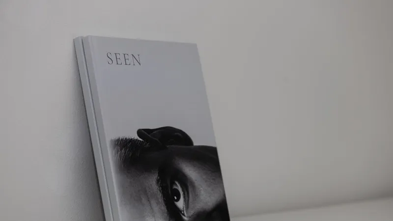 Two copies of the photo book SEEN leaning upright against a white wall, the cover showing an eye in a black-and-white portrait