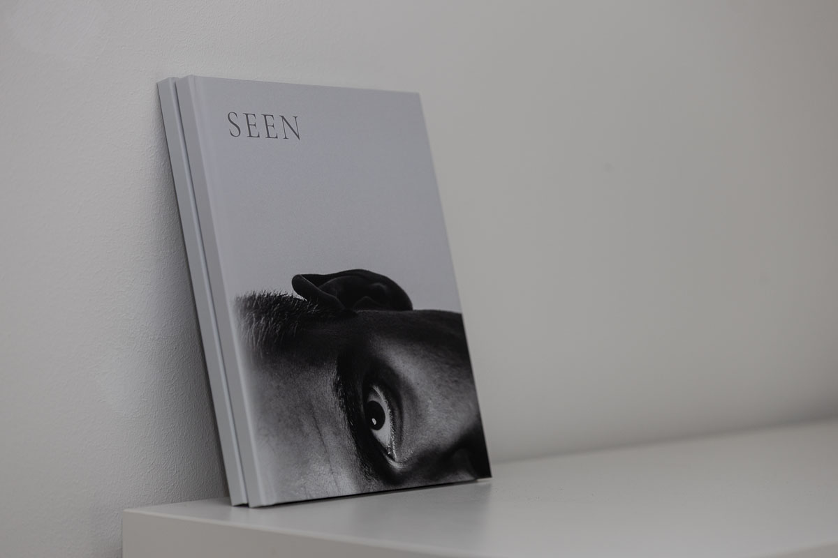 Zwei Exemplare des Fotobuchs SEEN lehnen aufrecht an einer weißen Wand, auf dem Cover ein Auge in einem Schwarz-Weiß-Porträt