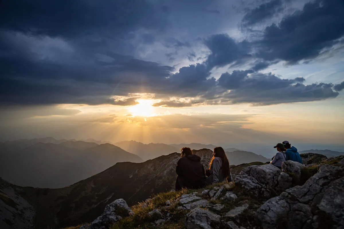 Several people sitting on a rocky mountain summit, looking out at a dramatic cloud formation and sunset.
