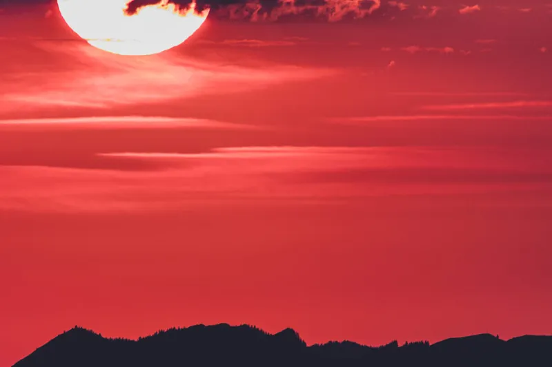 Half the sun glowing through breaking clouds above a dark mountain ridge, sky burning red.