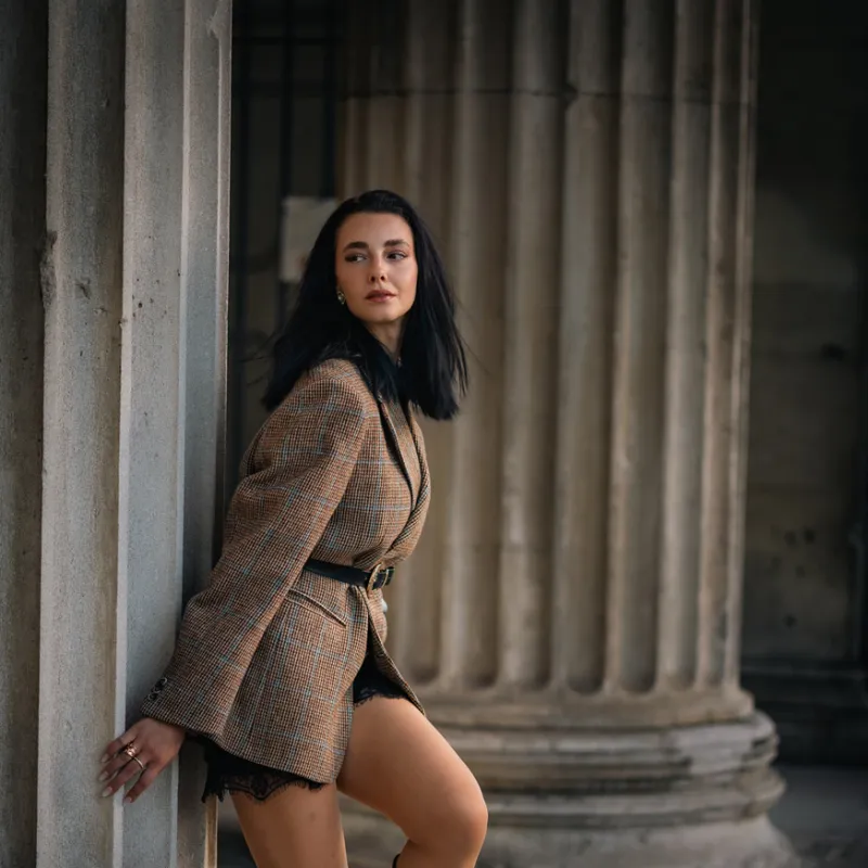 Monique in a tweed blazer and brown suede boots leaning against a stone column, looking over her shoulder