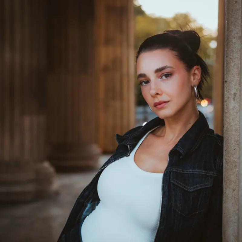 Monique in a denim jacket leaning against a column, looking into the camera, more columns blurred behind