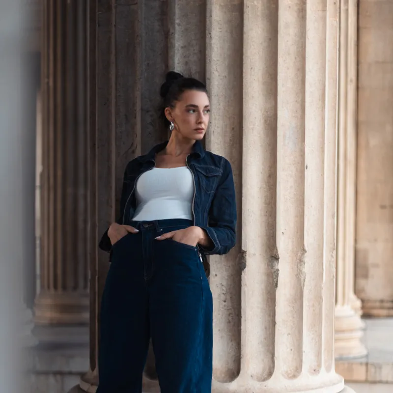 Monique in a denim jacket and wide jeans leaning against a column base, hands in her pockets