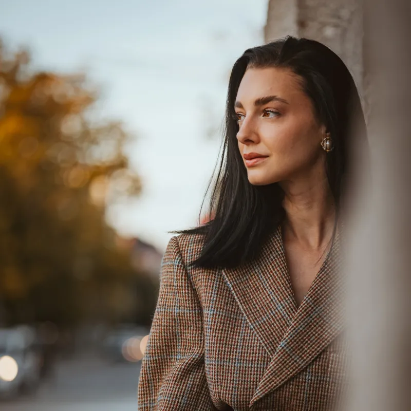 Monique in a tweed blazer with a gold belt buckle, looking to the side, a blurred street behind her