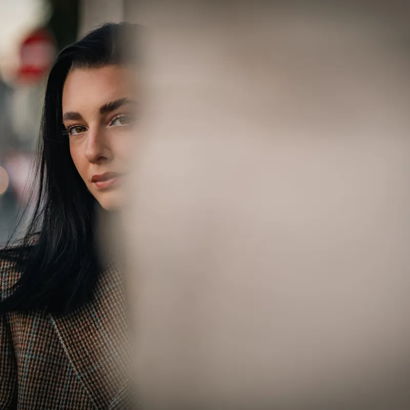 Half of Monique's face behind a blurred column in the foreground, tweed blazer, street lights out of focus