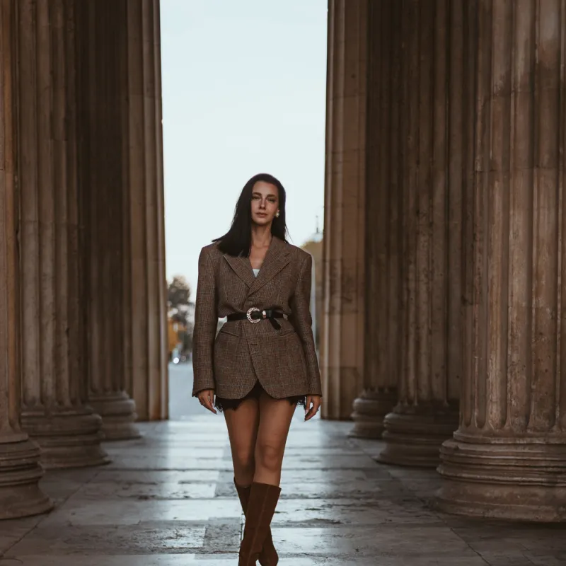 Monique walking toward the camera between large stone columns in a belted tweed blazer and brown suede boots