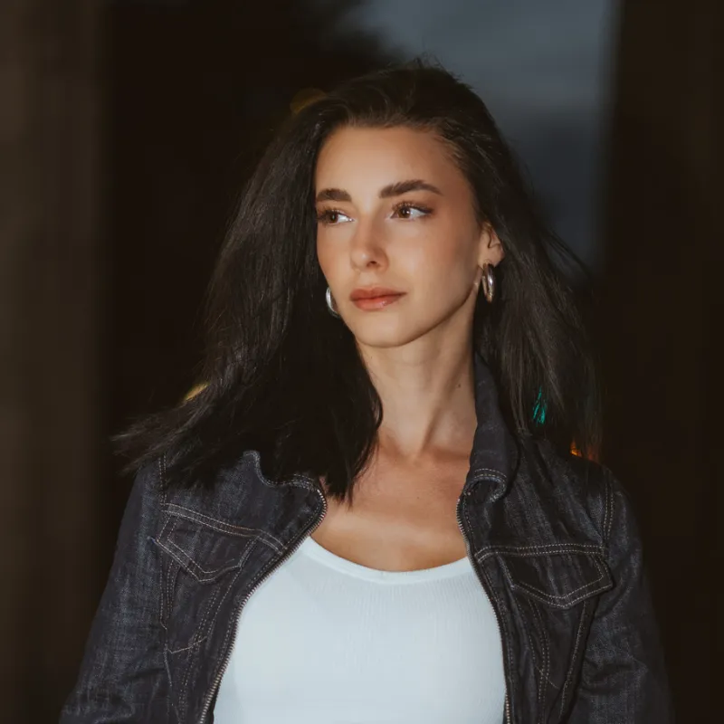 Monique in a denim jacket and white top, hands in her pockets, in front of a dark background