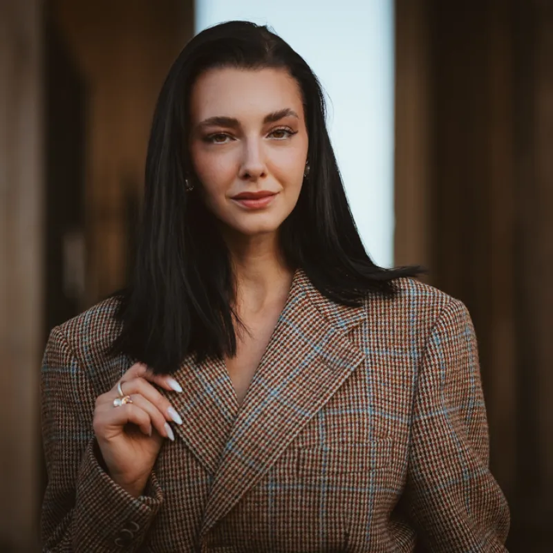 Monique in a checked tweed blazer, one hand at her hair, in front of blurred stone columns