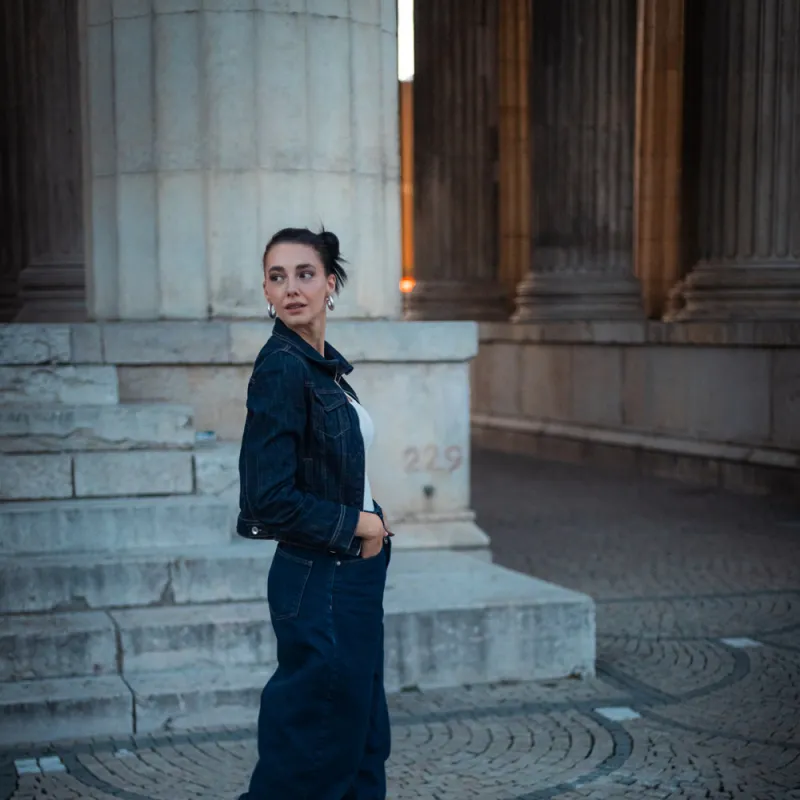 Monique in a denim jacket and wide jeans walking across cobblestones, looking back over her shoulder