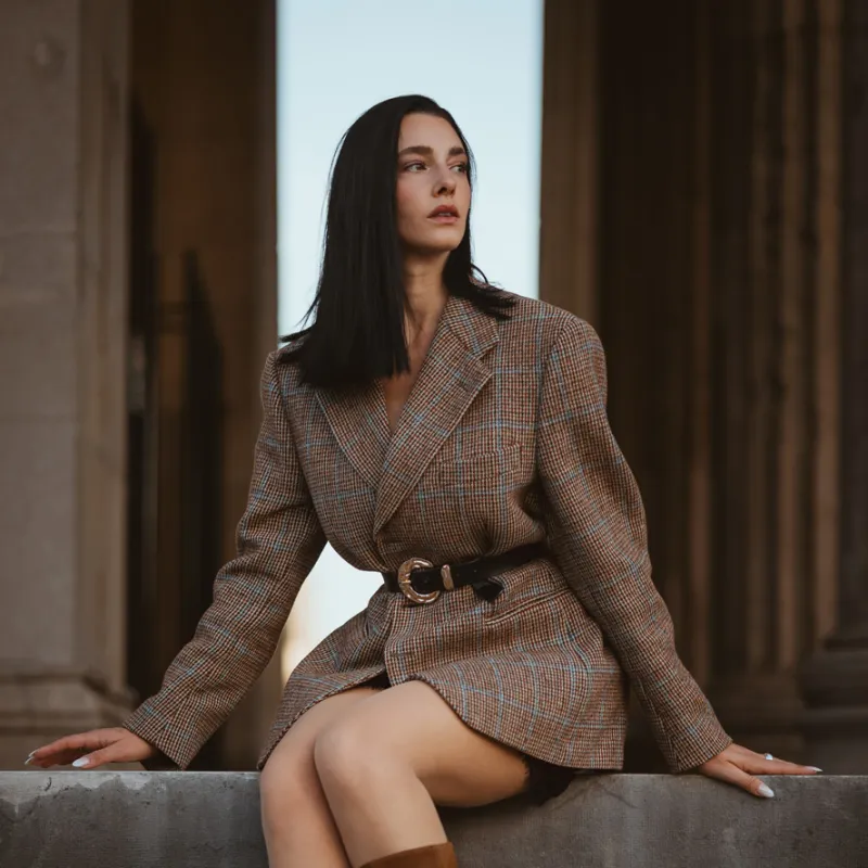 Monique sitting on a stone ledge in a tweed blazer and brown suede boots, columns behind her