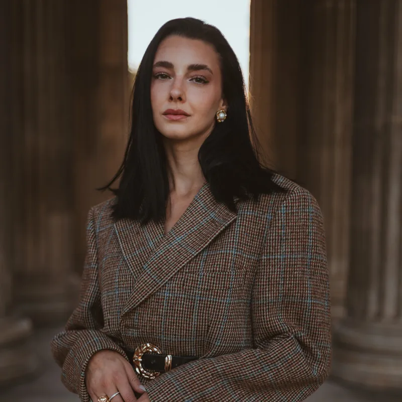 Monique in a tweed blazer with a gold belt buckle, hands folded in front, columns behind her