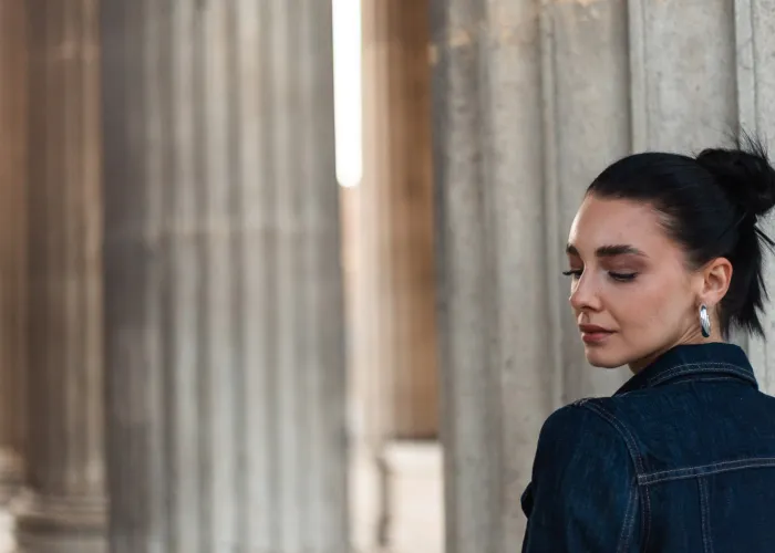 Monique in profile with her hair in a bun, eyes lowered, in a denim jacket next to a row of columns