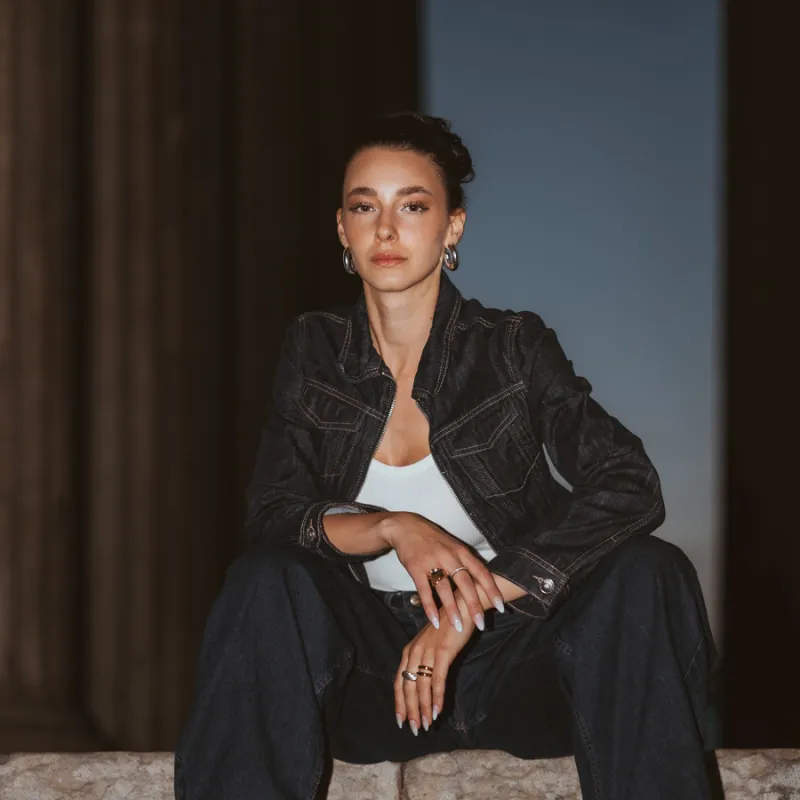 Monique sitting on stone steps in a denim outfit, one knee raised, looking to the side at dusk