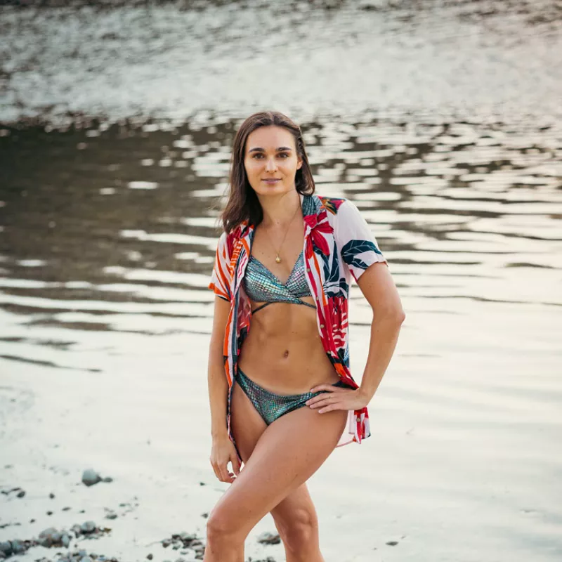 Eine Frau im glitzernden Bikini und offenem Blumenhemd steht am Flussufer.