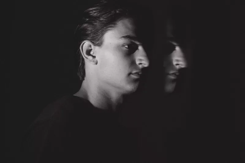 Black and white portrait of a young man in profile against a near-black background. A faint reflection appears beside him, creating a sense of duality and quiet tension.