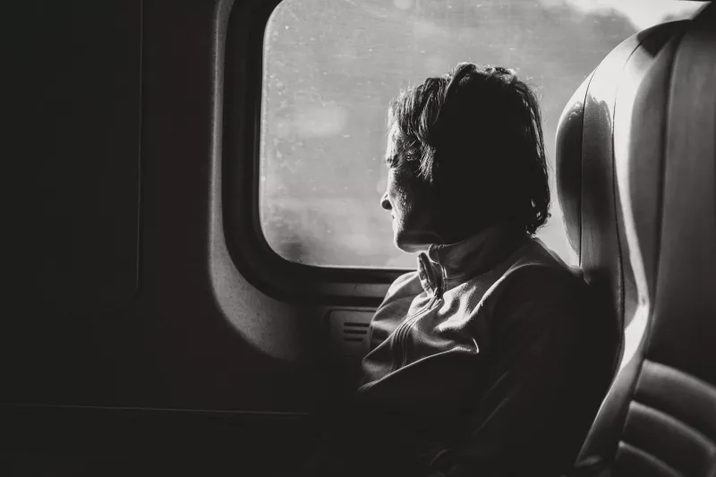 Black and white photograph of an elderly woman sitting by a train window, face in profile, gazing outside. Side light from the window creates strong contrast against the dark interior.